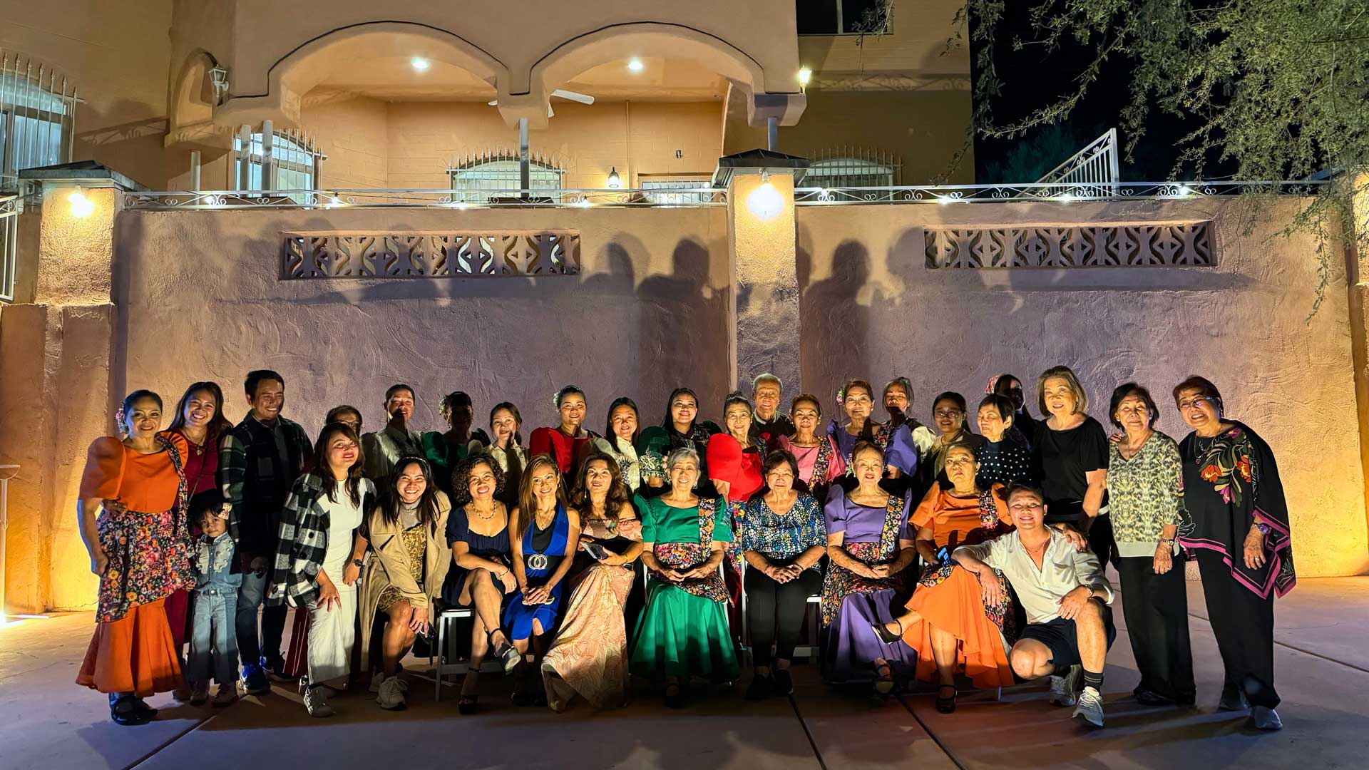 A large group of smiling people, mostly women, pose for a night photo outdoors in front of an illuminated building.