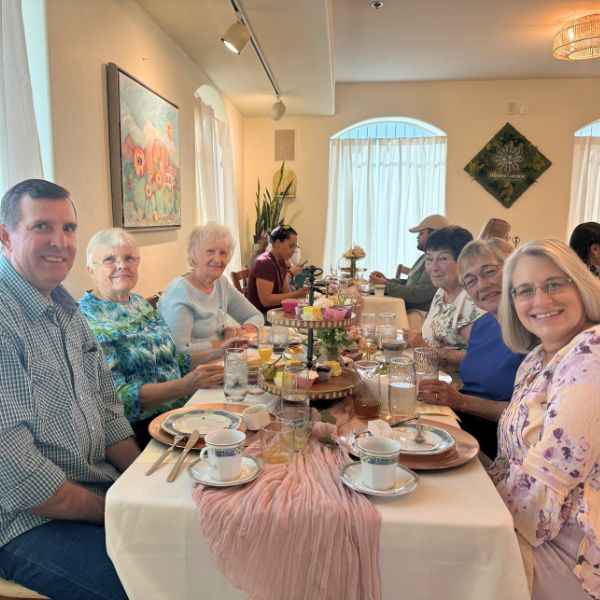 A diverse group enjoys a bright meal, smiling at the camera, with desserts and tea on the table.