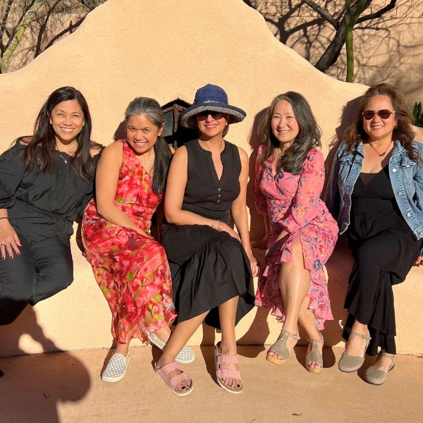 Five women, including the Himaya Garden owners, sit side-by-side on a light-colored, textured wall outdoors, smiling.