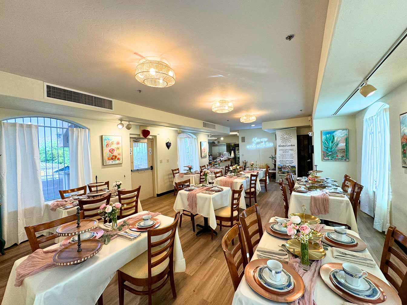 A brightly lit indoor dining area at Himaya Garden features multiple tables set for tea, with white tablecloths, pink runners, and elegant place settings.