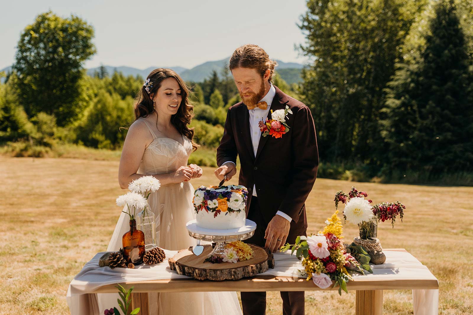 A smiling bride in a light-colored dress watches as her groom, in a dark suit with a floral boutonniere, cuts a vibrantly decorated wedding cake outdoors.