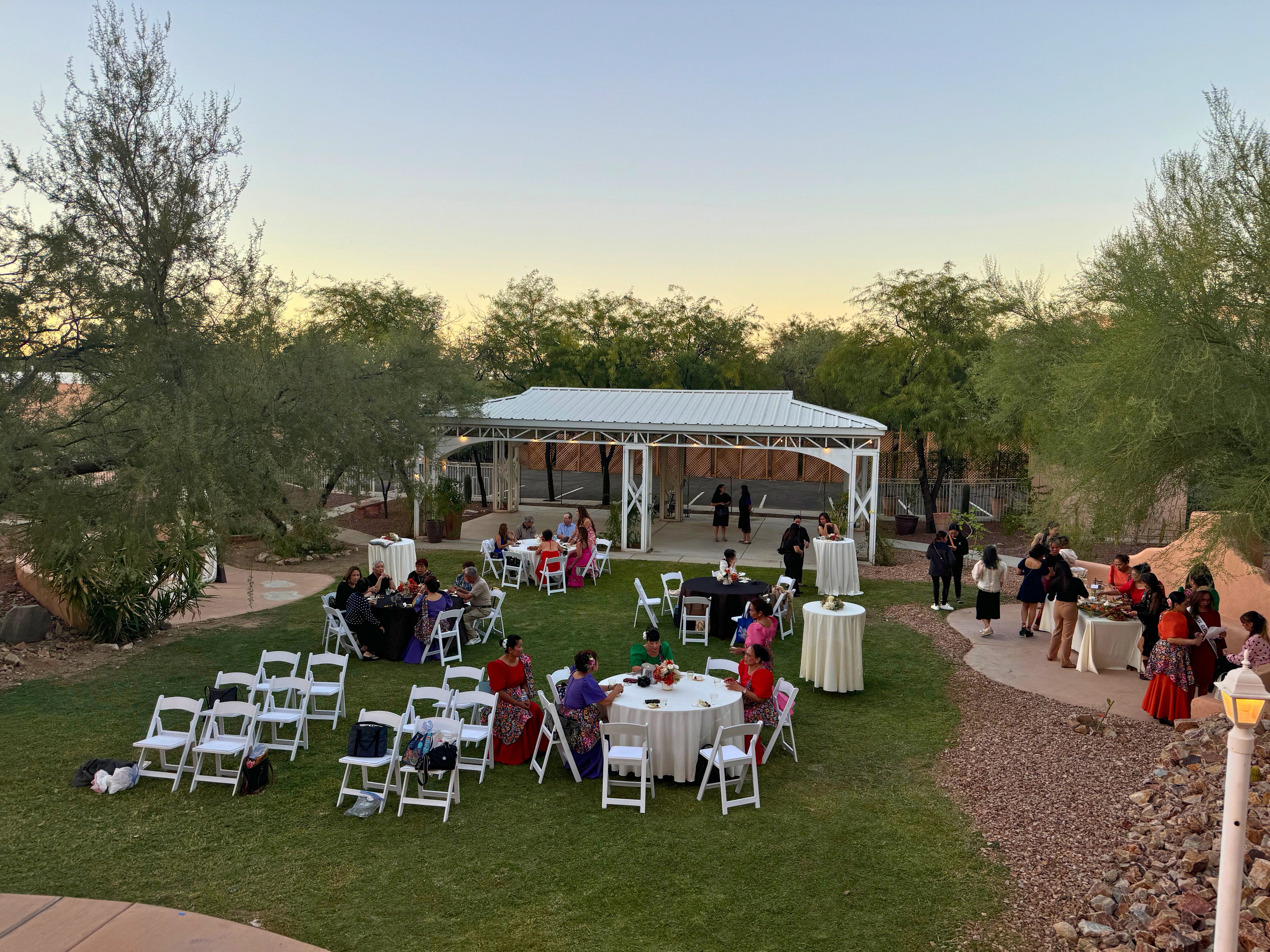 People mingle on the green lawn of Himaya Garden's outdoor area during an event. White chairs, round tables, and a large white pavilion are visible.