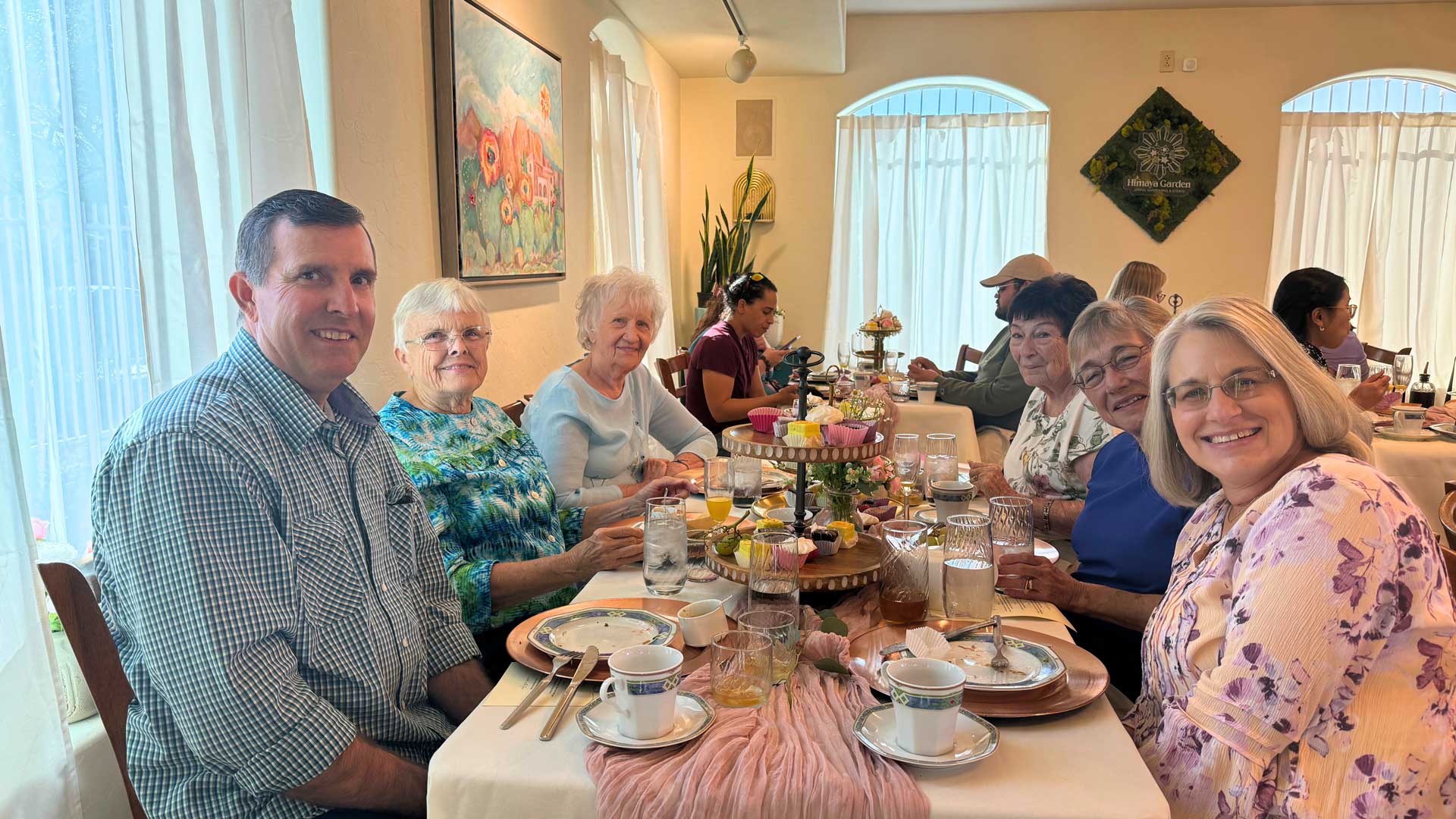 A diverse group enjoys a bright meal, smiling at the camera, with desserts and tea on the table.